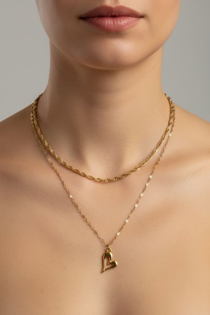 Model wearing layered gold necklaces including a double chain with a small heart pendant