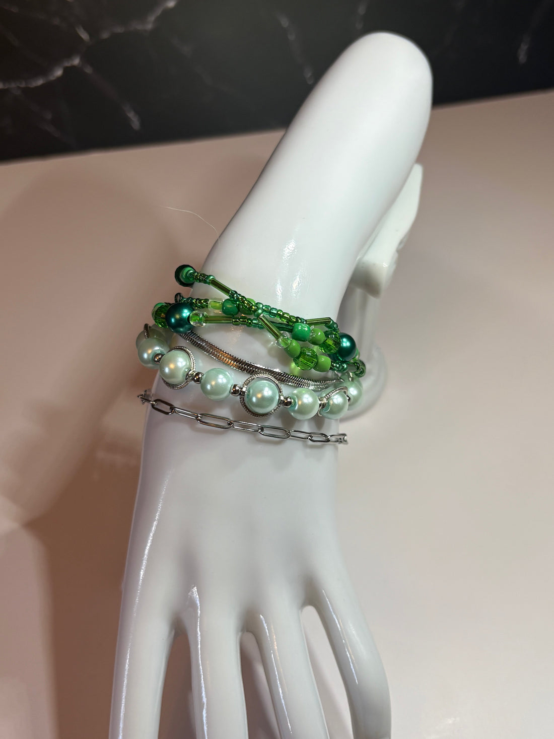 A stacked bracelet set in green shades displayed on a white jewellery hand, featuring beaded, pearl, chain and wire bracelets layered together