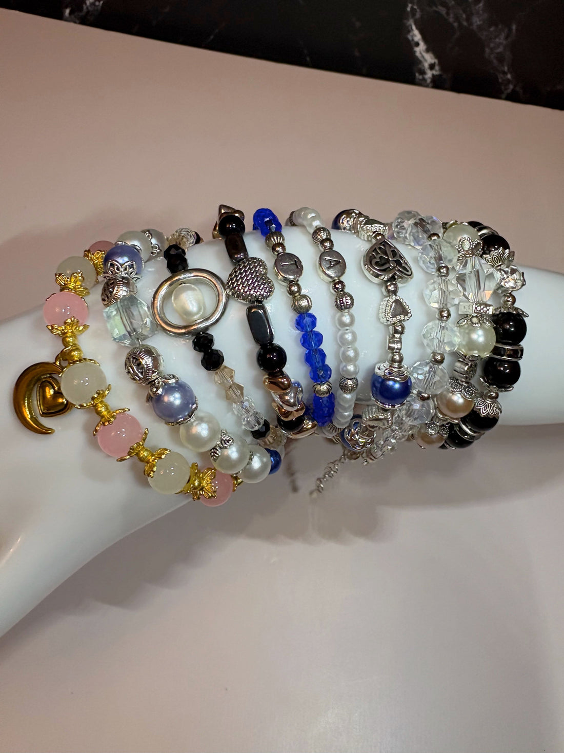 A collection of beaded bracelets displayed on a person's wrist, featuring a variety of styles including black and silver, white pearl, blue, and gold tones, with small pendant charms.