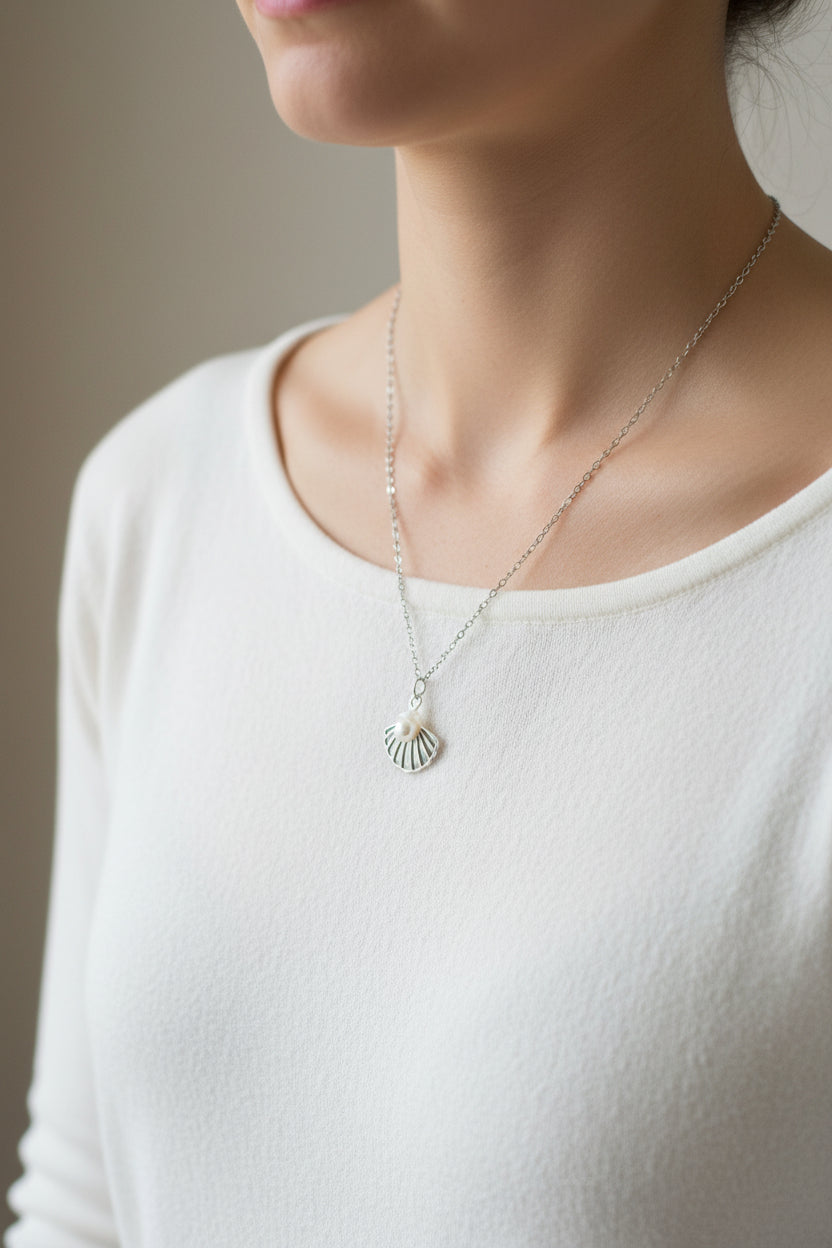 A silver pendant necklace with a small fan or scallop-shaped charm, displayed on a model wearing a white top