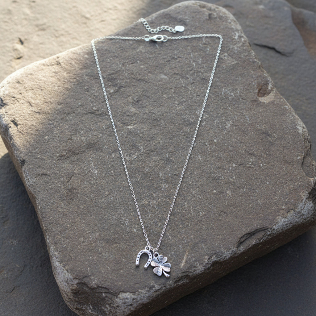 A silver-toned chain necklace with a small charm set, including a four-leaf clover and a horseshoe pendant, displayed on a light beige background next to a penny for scale.