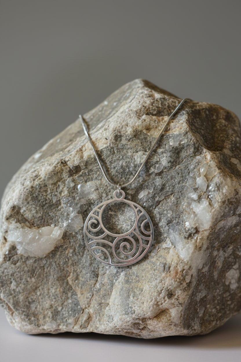 A silver circular pendant necklace with an intricate scroll cutout pattern, displayed on a large stone.