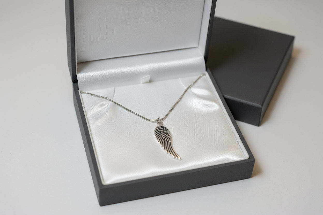 A silver wing pendant necklace displayed inside an open dark gray gift box with a white satin lining, alongside a closed matching box on a light gray background.