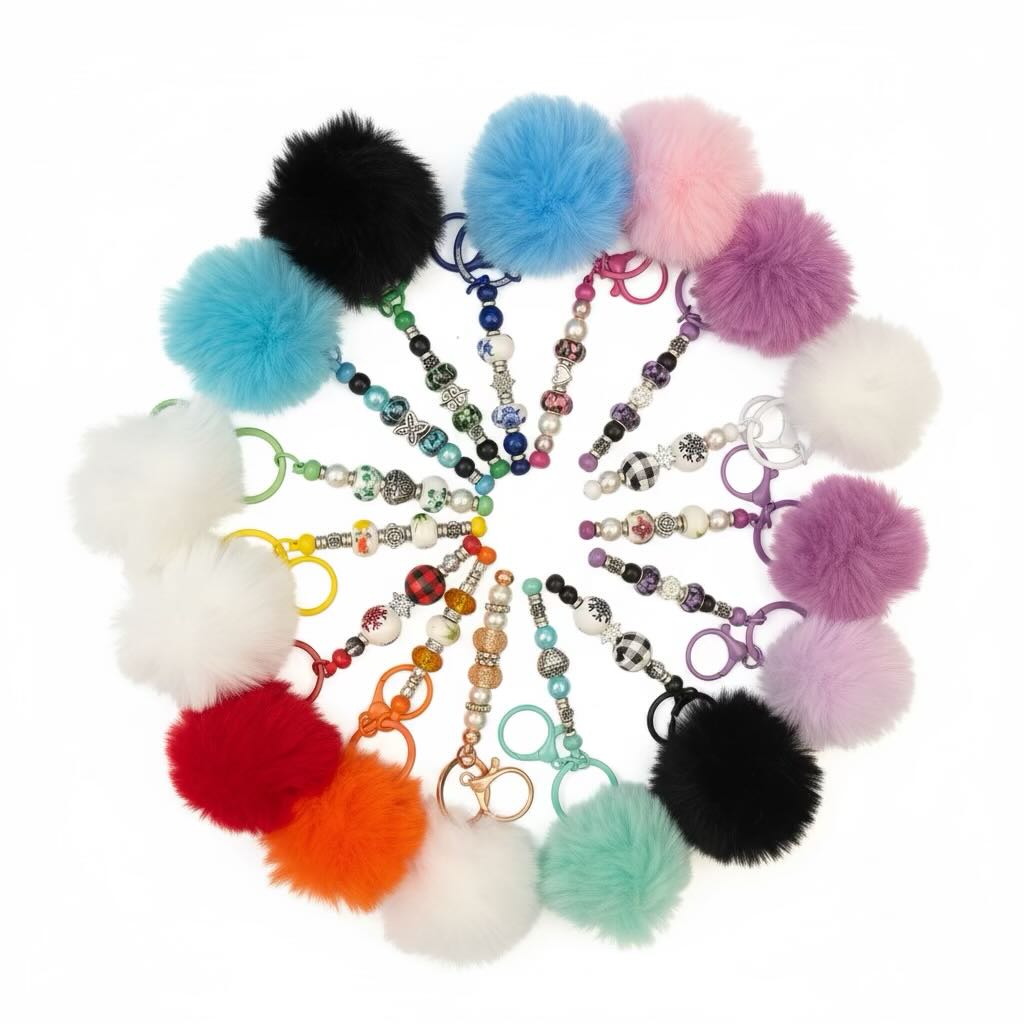 A collection of colorful faux fur pom-pom keychains arranged in a circular pattern on a white background. Each keychain features a fluffy pom-pom in various colors including black, blue, pink, purple, white, green, yellow, orange, and mint, attached to a beaded key ring.