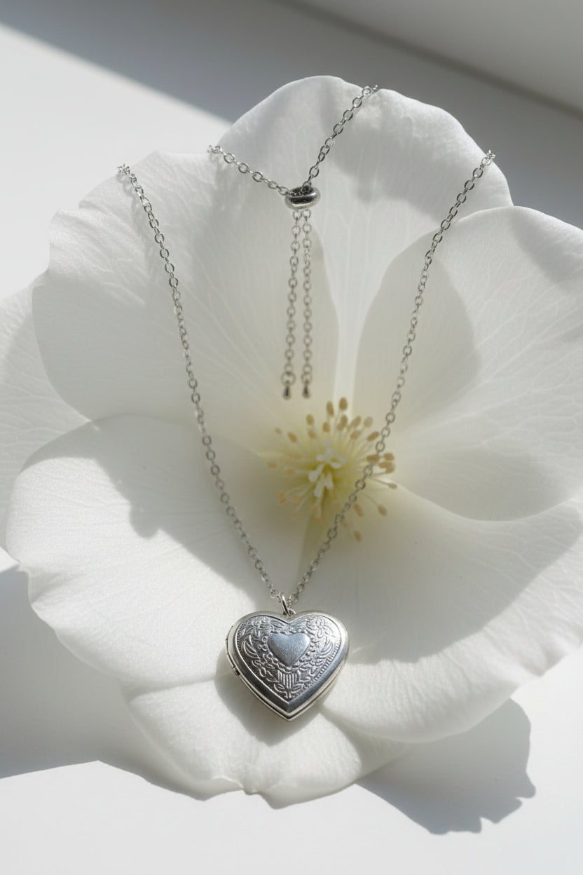 A silver heart-shaped pendant necklace with a textured outer border and a smaller silver heart in the center, displayed on a large white flower.