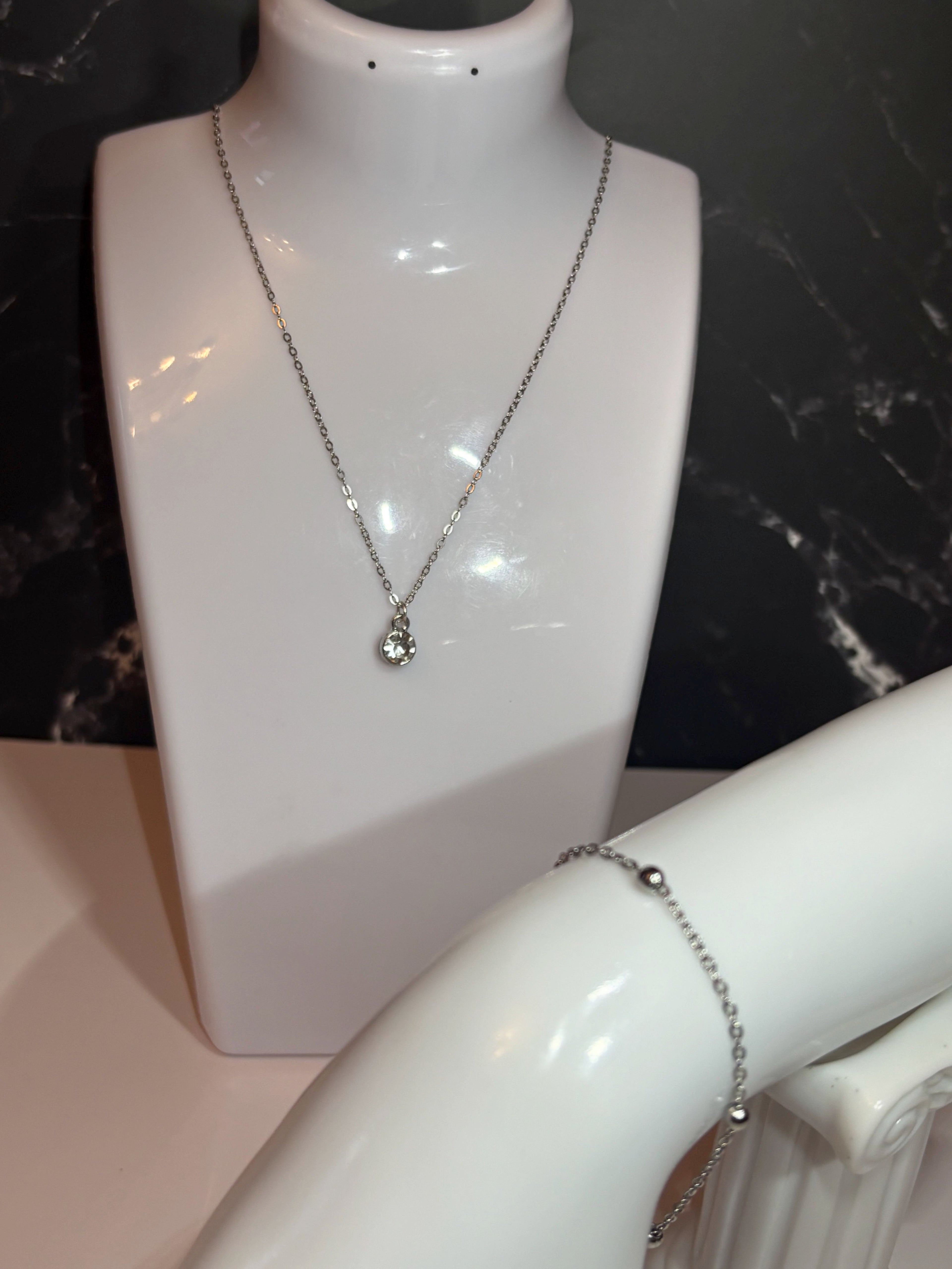 A silver-toned necklace with a small diamond birthstone pendant displayed on a white jewelry bust, with a bracelet partially visible on another arm stand against a dark marble background.