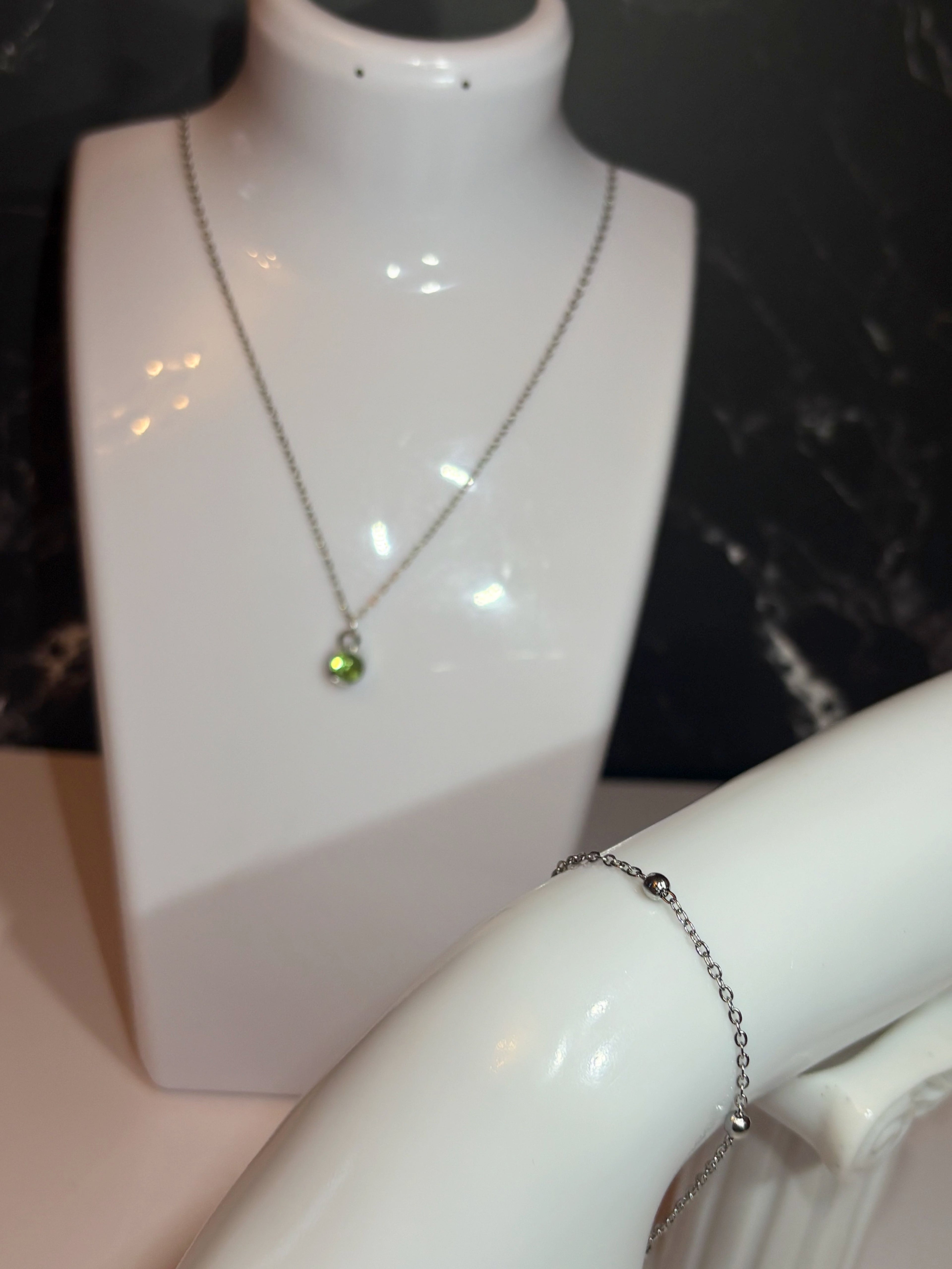 A silver-toned chain necklace with a small green birthstone pendant displayed on a white jewelry bust, alongside a silver ball chain bracelet on a curved white arm.