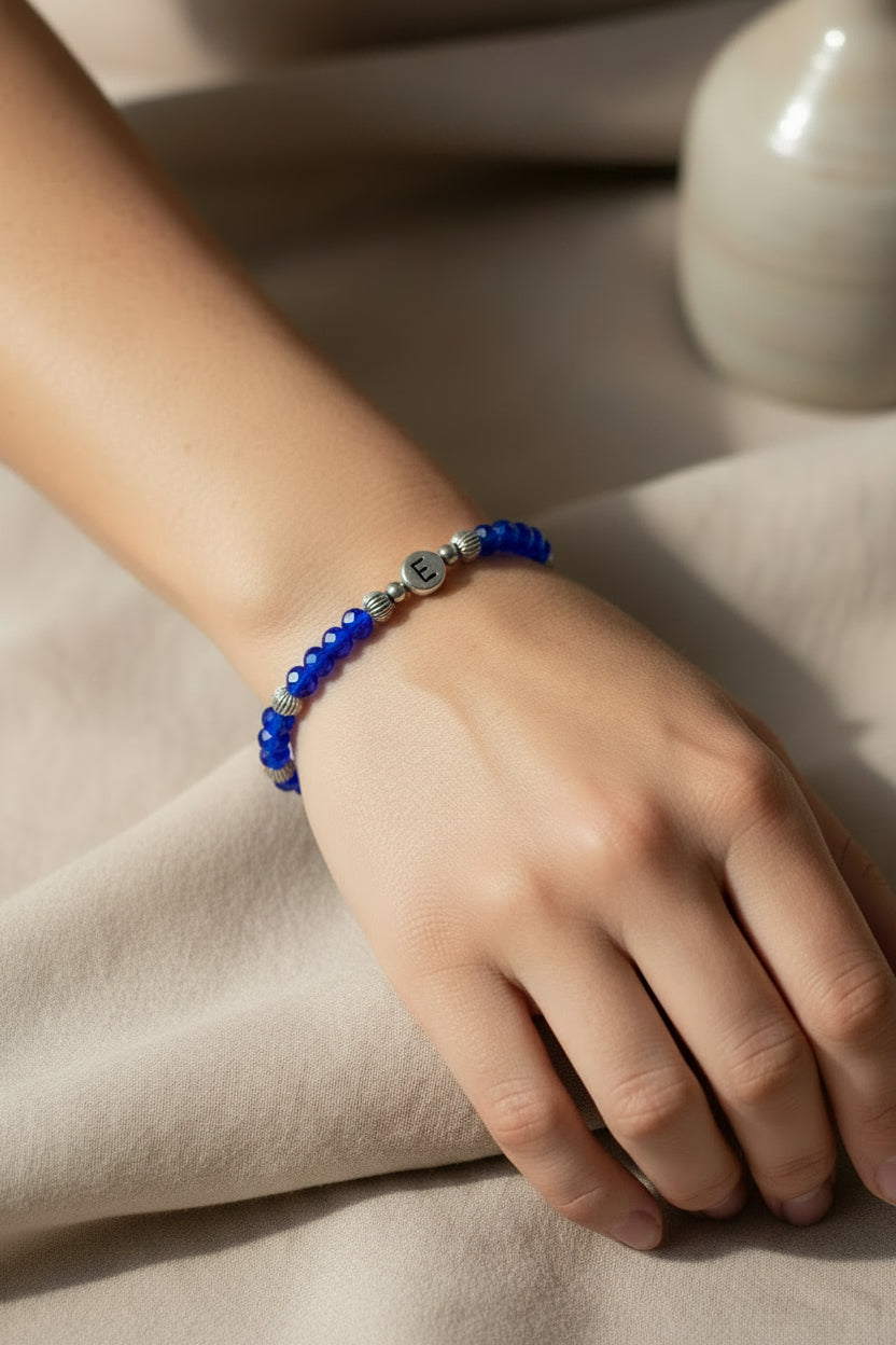 A blue beaded bracelet with silver spacer beads and a central silver letter charm spelling 'E', displayed on a white mannequin arm.