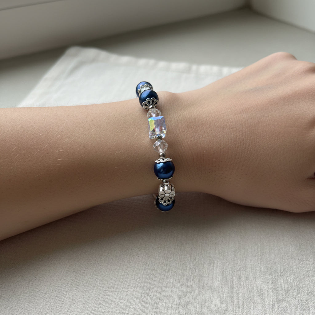 A beaded bracelet with dark royal blue glass pearls, clear faceted crystal beads, and silver-tone metal flower charms, arranged in a circle on a plain light background.