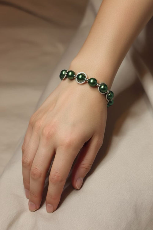 A close-up of a person's hand and wrist wearing a bracelet made of dark green round pearls separated by small silver spacer rings, resting on a soft beige fabric surface.