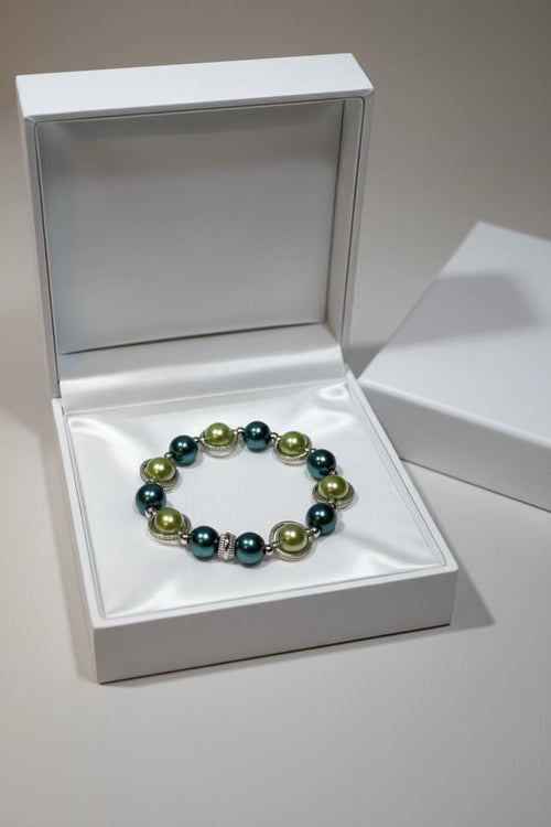 A beaded bracelet with dark teal and sage green pearls, displayed inside an open white jewelry box.