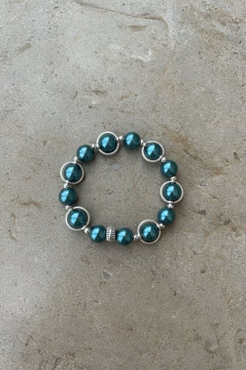 A beaded stretch bracelet with teal-green round pearls set in silver-tone metal, featuring a small textured spacer bead, displayed on a neutral gray stone surface.