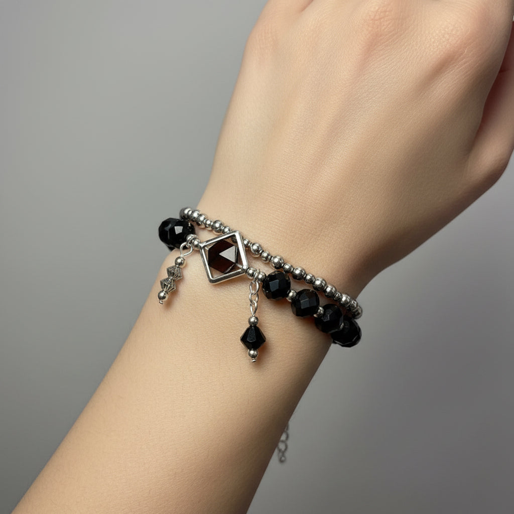 A gothic-style beaded bracelet with black faceted beads, a silver-toned chain, and a central dark gemstone charm, displayed on a white mannequin hand.