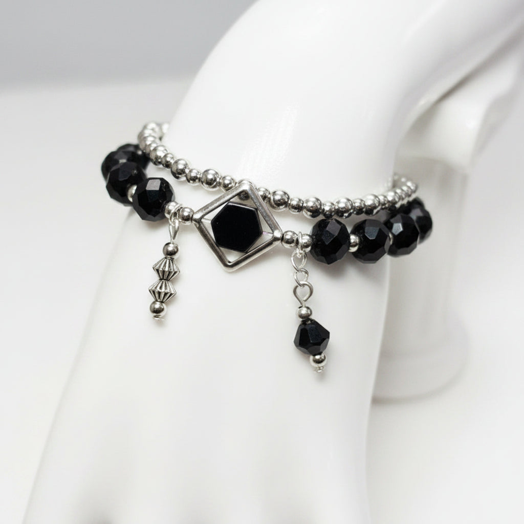 A gothic-style double-strand bracelet displayed on a white mannequin hand. The bracelet features silver-toned beads, faceted black beads, a central silver diamond-shaped charm with a dark red gemstone, and dangling silver and black charms.