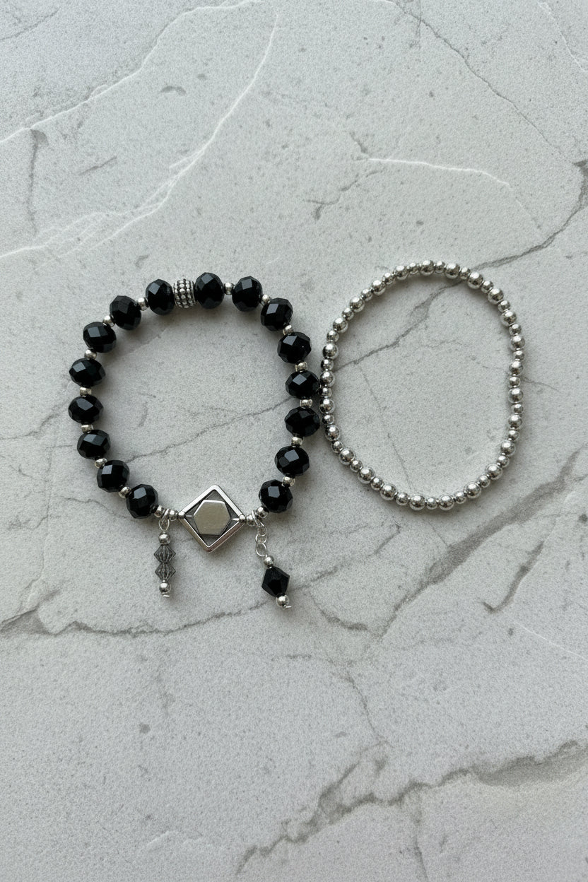 Two beaded bracelets displayed on a plain light background: one bracelet features black faceted beads with a silver-toned diamond-shaped charm and dangling beads, while the other is a simple silver-toned beaded bracelet.