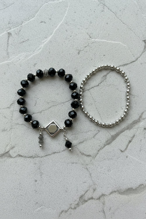 Two beaded bracelets displayed on a plain light background: one bracelet features black faceted beads with a silver-toned diamond-shaped charm and dangling beads, while the other is a simple silver-toned beaded bracelet.