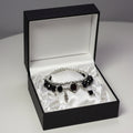 A gothic-style double-strand bracelet displayed on a white mannequin hand. The bracelet features silver-toned beads, faceted black beads, a central silver diamond-shaped charm with a dark red gemstone, and dangling silver and black charms.