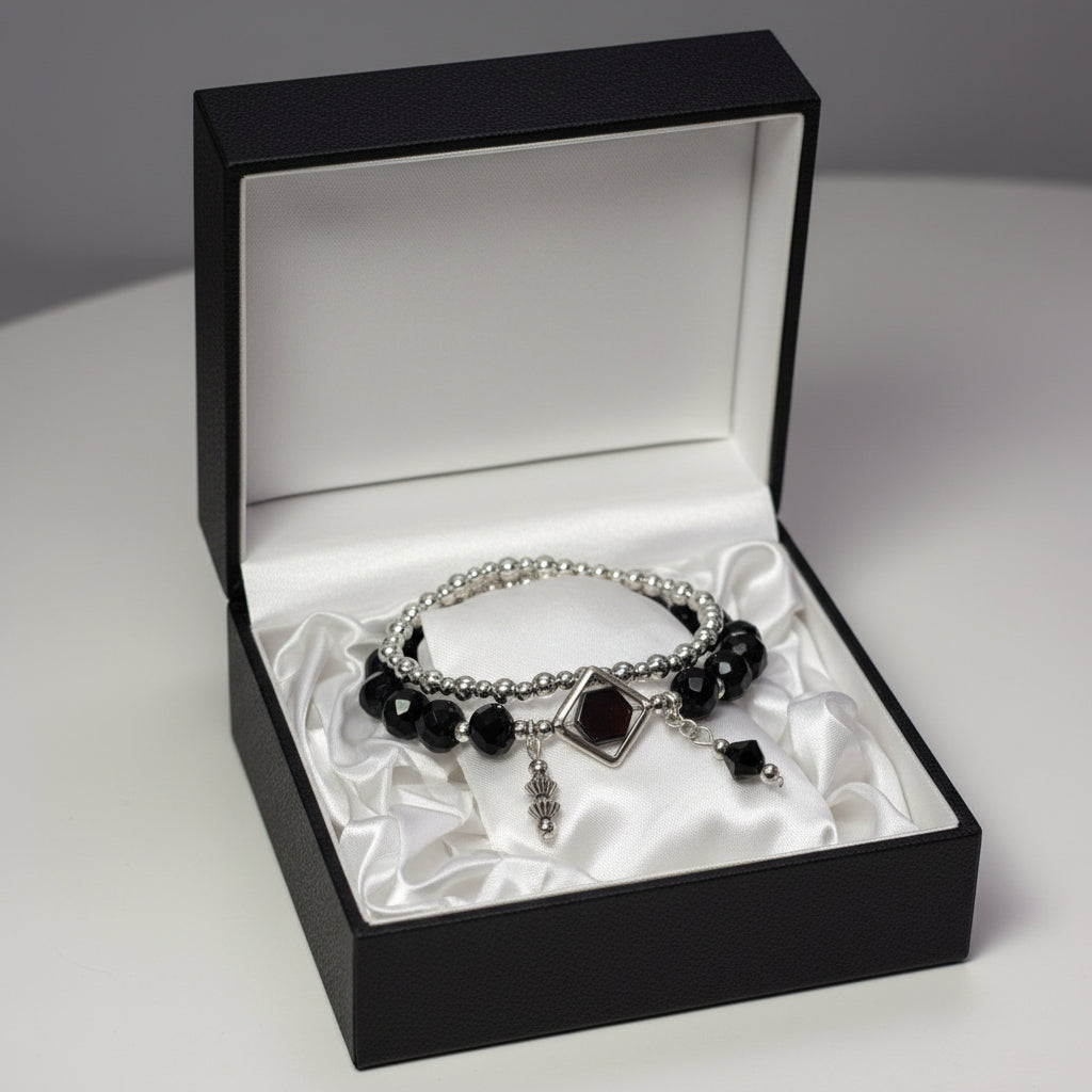 A gothic-style double-strand bracelet displayed on a white mannequin hand. The bracelet features silver-toned beads, faceted black beads, a central silver diamond-shaped charm with a dark red gemstone, and dangling silver and black charms.