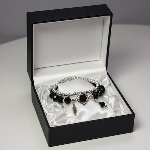 A gothic-style double-strand bracelet displayed on a white mannequin hand. The bracelet features silver-toned beads, faceted black beads, a central silver diamond-shaped charm with a dark red gemstone, and dangling silver and black charms.