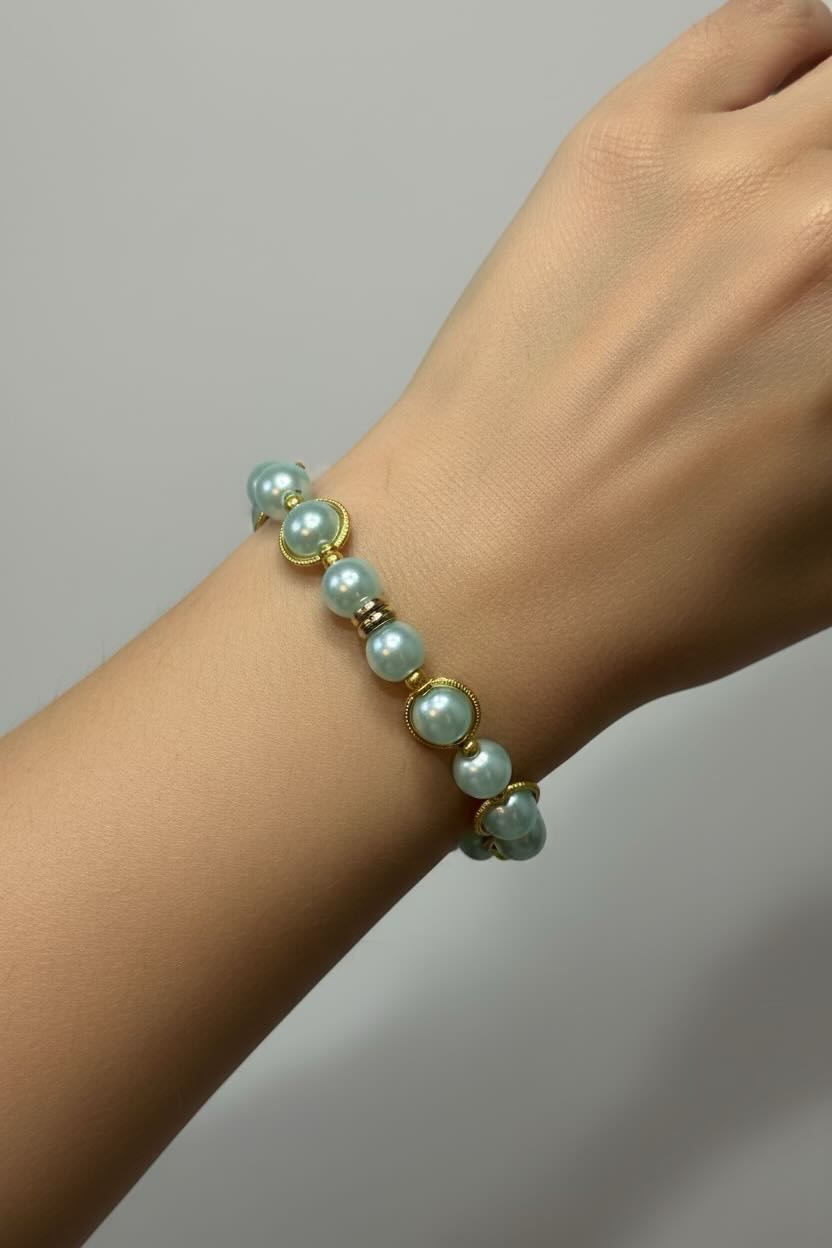 A close-up of a person's wrist wearing a pearl bracelet made of aqua blue-pearls with gold-toned spacers and settings, against a plain light gray background.
