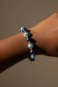 A close-up of a beaded bracelet worn on a person's wrist. The bracelet features alternating dark royal blue beads and dark baby blue glass pearls, separated by small gold spacer rings, set against a solid warm brown background.