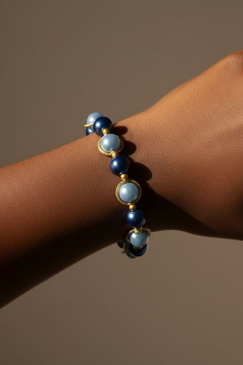 A close-up of a beaded bracelet worn on a person's wrist. The bracelet features alternating dark royal blue beads and dark baby blue glass pearls, separated by small gold spacer rings, set against a solid warm brown background.