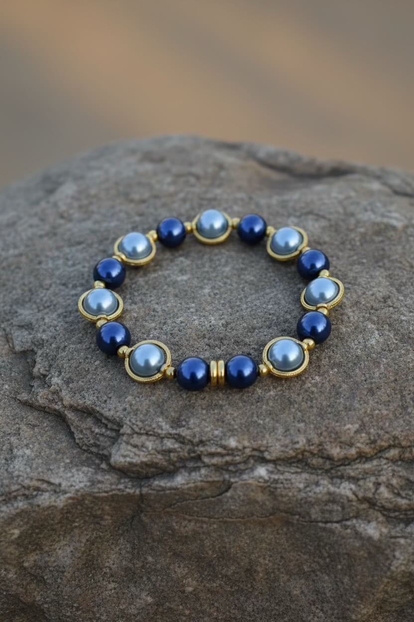 A beaded stretch bracelet with alternating dark royal blue and dark baby blue round glass pearls, accented by gold spacer beads, displayed on a textured gray rock.