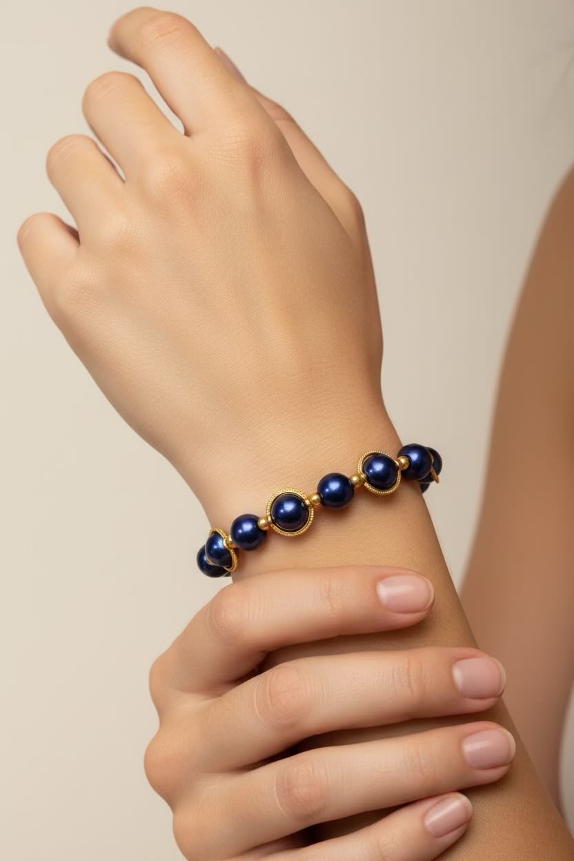 A close-up of a person's wrist wearing a beaded bracelet with dark royal blue round glass pearls and gold-toned metal links, against a soft beige background.