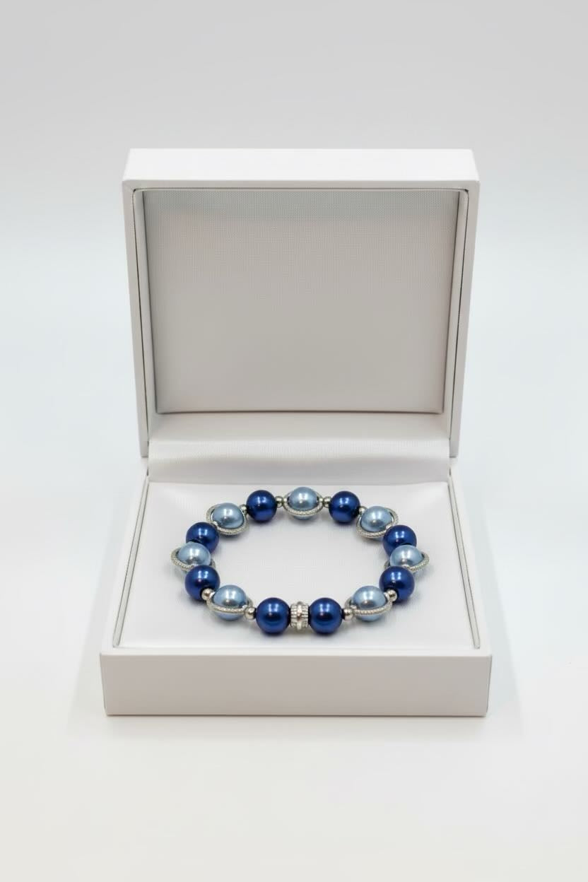 A beaded stretch bracelet with alternating deep blue and light blue pearls, accented with small silver spacer beads, displayed inside an open white jewelry box on a white background.