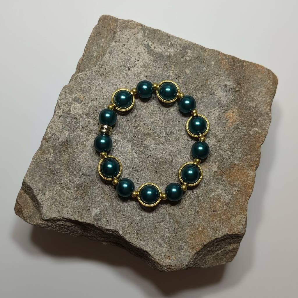 A beaded bracelet with teal-green round glass pearls and gold-toned spacer rings, displayed on a rough gray stone against a plain light background.