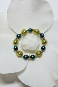 A beaded stretch bracelet with alternating teal green and sage green pearls in gold-toned bezel settings, displayed on a large white orchid flower.