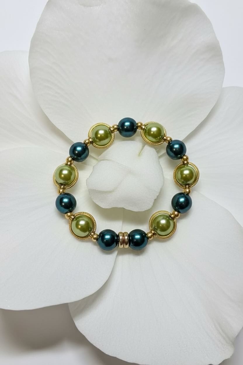 A beaded stretch bracelet with alternating teal green and sage green pearls in gold-toned bezel settings, displayed on a large white orchid flower.
