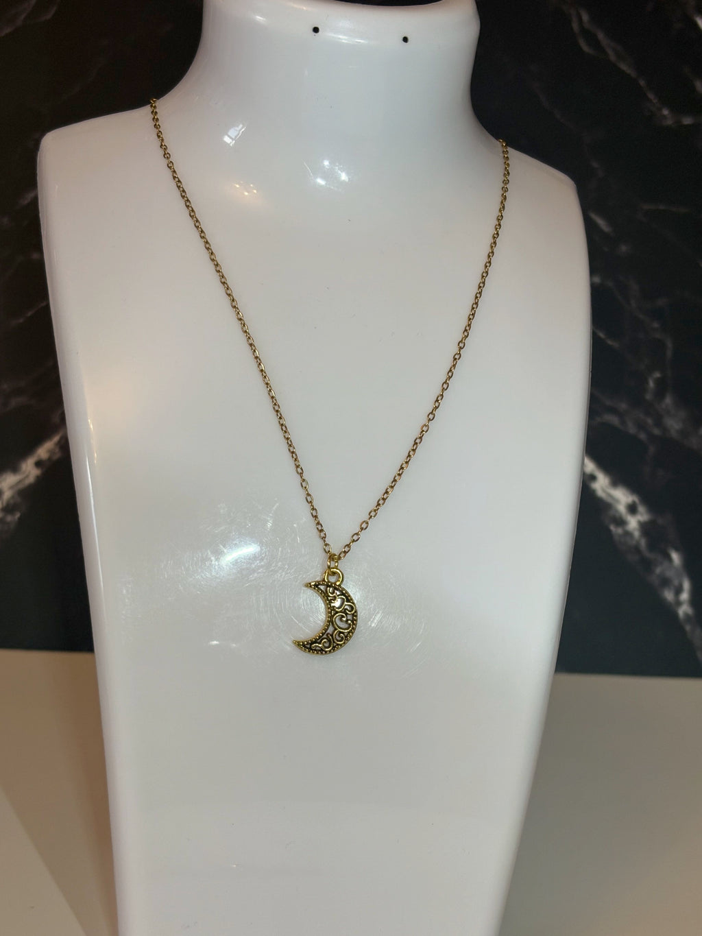 A gold crescent moon pendant necklace displayed on a white mannequin bust against a dark marble-patterned background.