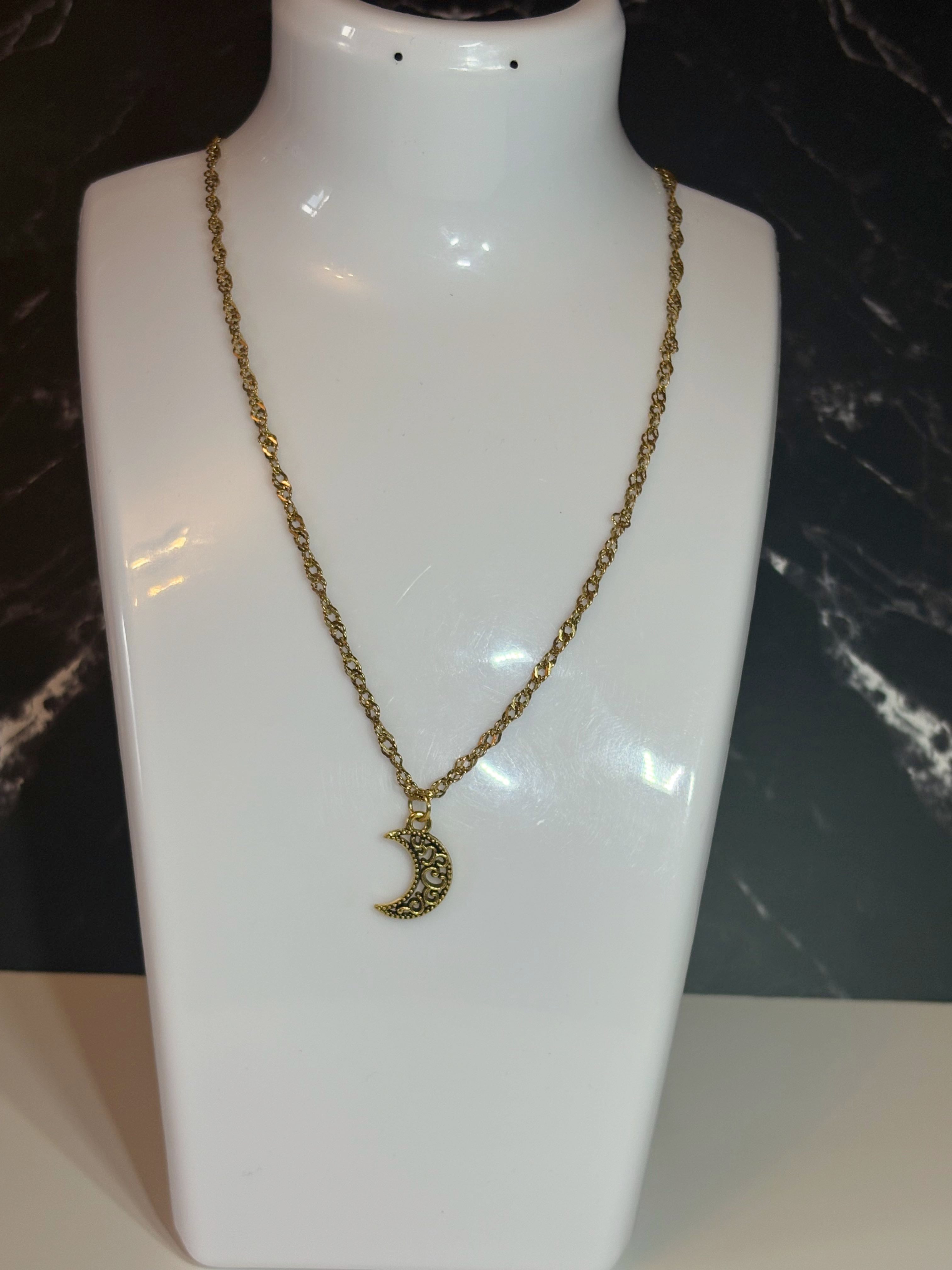 A gold-toned chain necklace with a crescent moon pendant, displayed on a glossy white jewelry bust against a dark marble-patterned background.