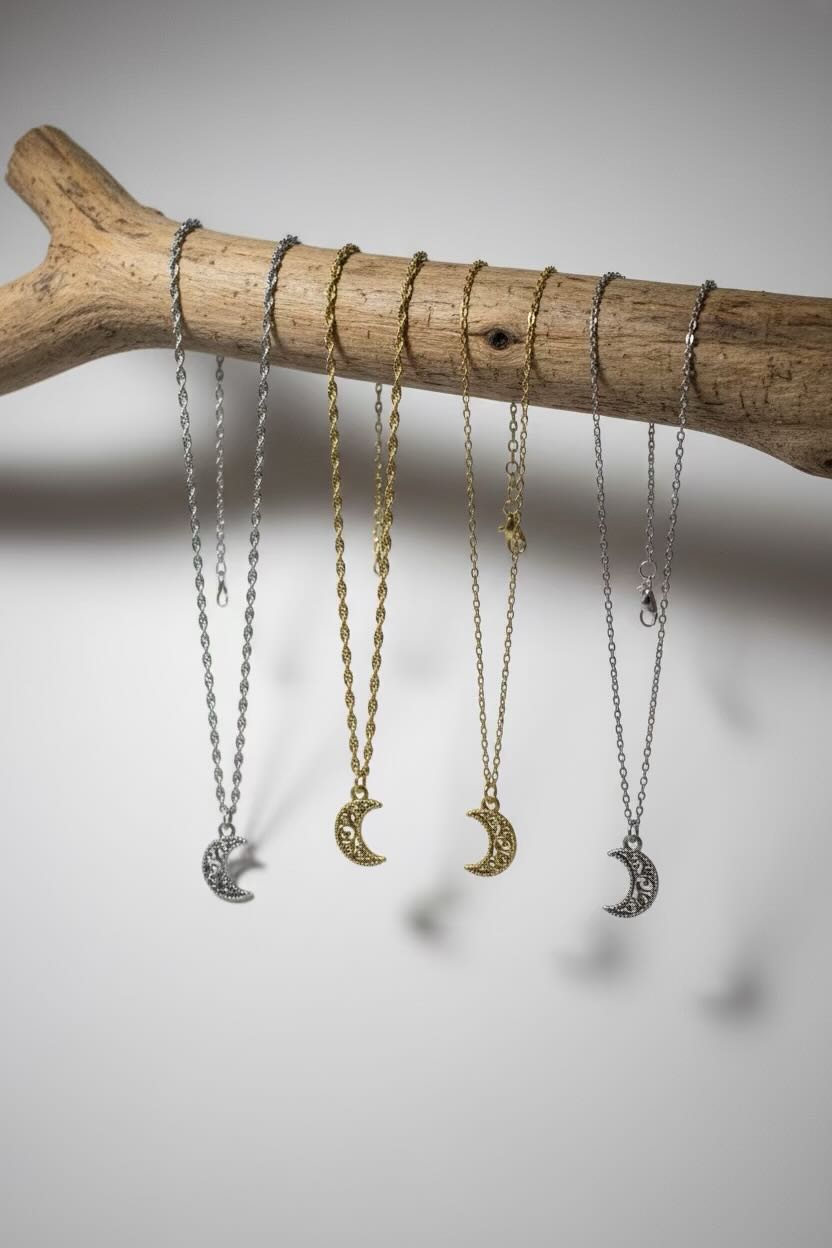 Four crescent moon pendant necklaces in silver and gold tones hang from a natural wood branch against a neutral gray background.