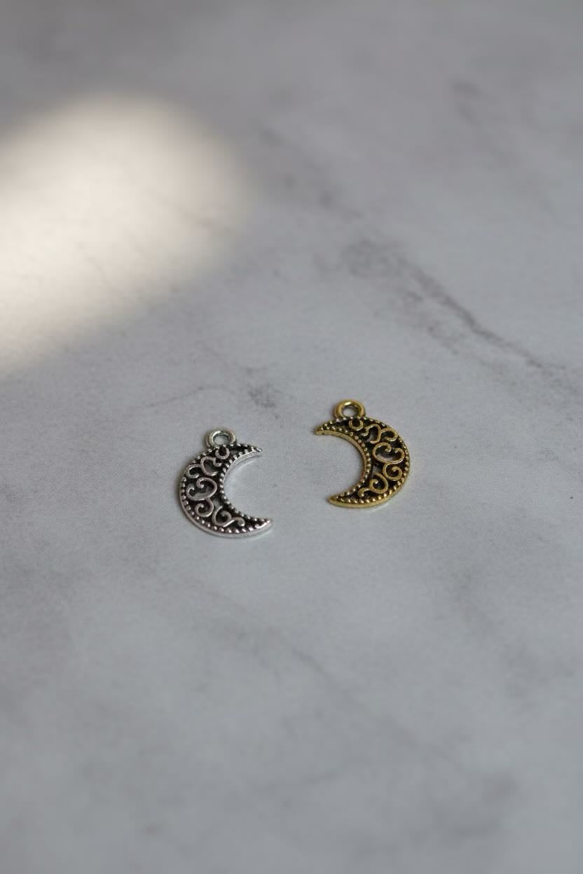 Two small crescent moon charms with ornate filigree detailing, one silver-toned and one gold-toned, displayed on a light gray marble surface.