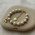 A gold-tone bracelet featuring white faux pearls, decorative gold spacer beads, a lobster clasp, an extender chain with a heart-shaped tag, and a gold-tone fleur-de-lis charm, displayed on a large stone.