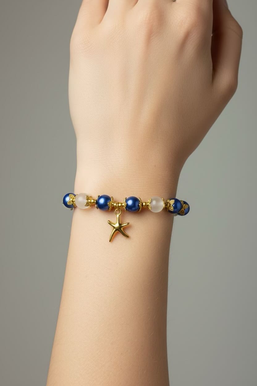 A close-up of a person's wrist wearing a beaded bracelet with blue and white beads, gold-toned details, and a small gold starfish charm, against a neutral gray background.