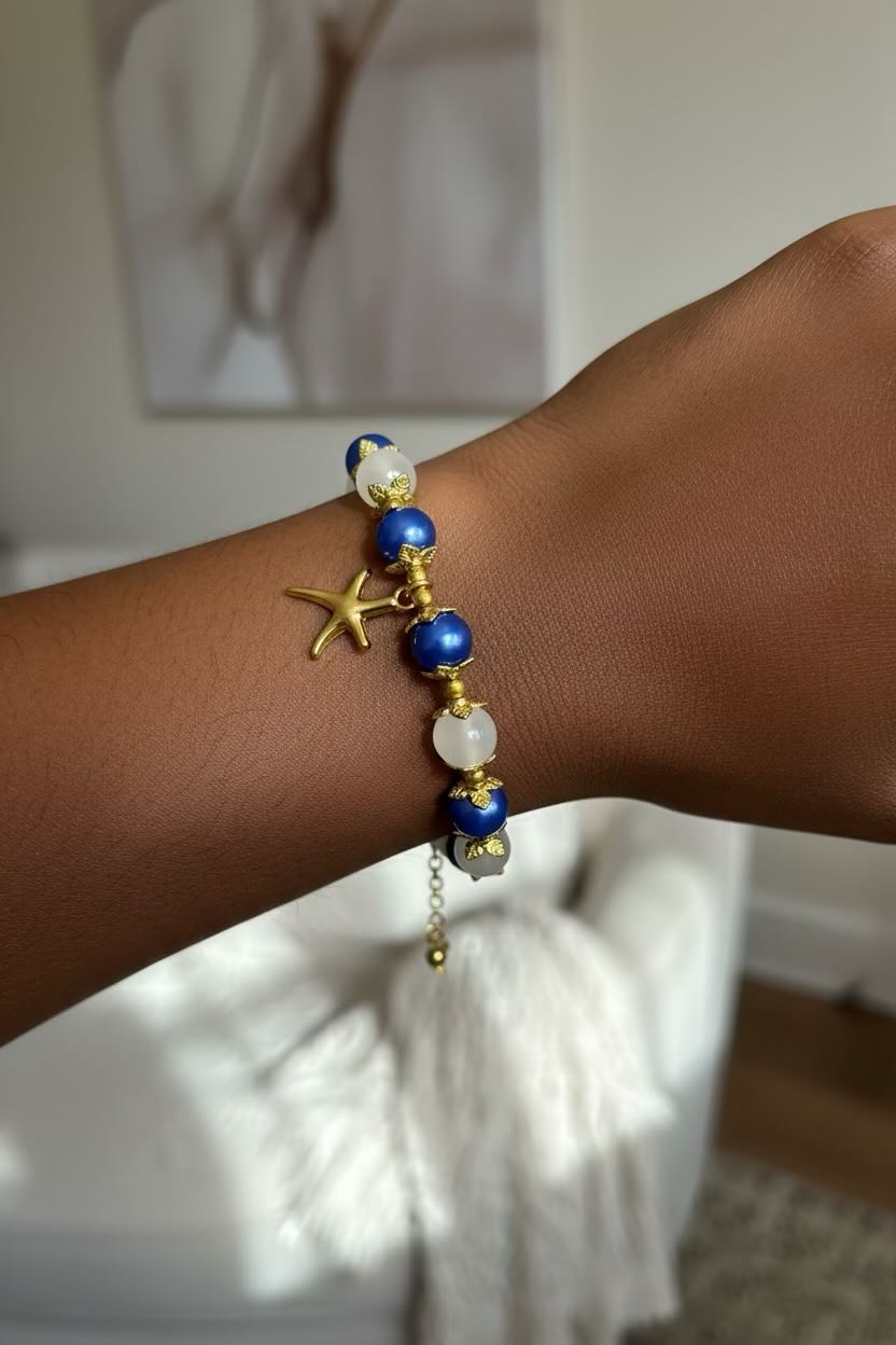 A close-up of a person's wrist wearing a beaded bracelet with blue beads, white pearl-like beads, gold-toned spacer details, a small gold starfish charm, and a dangling gold bead.