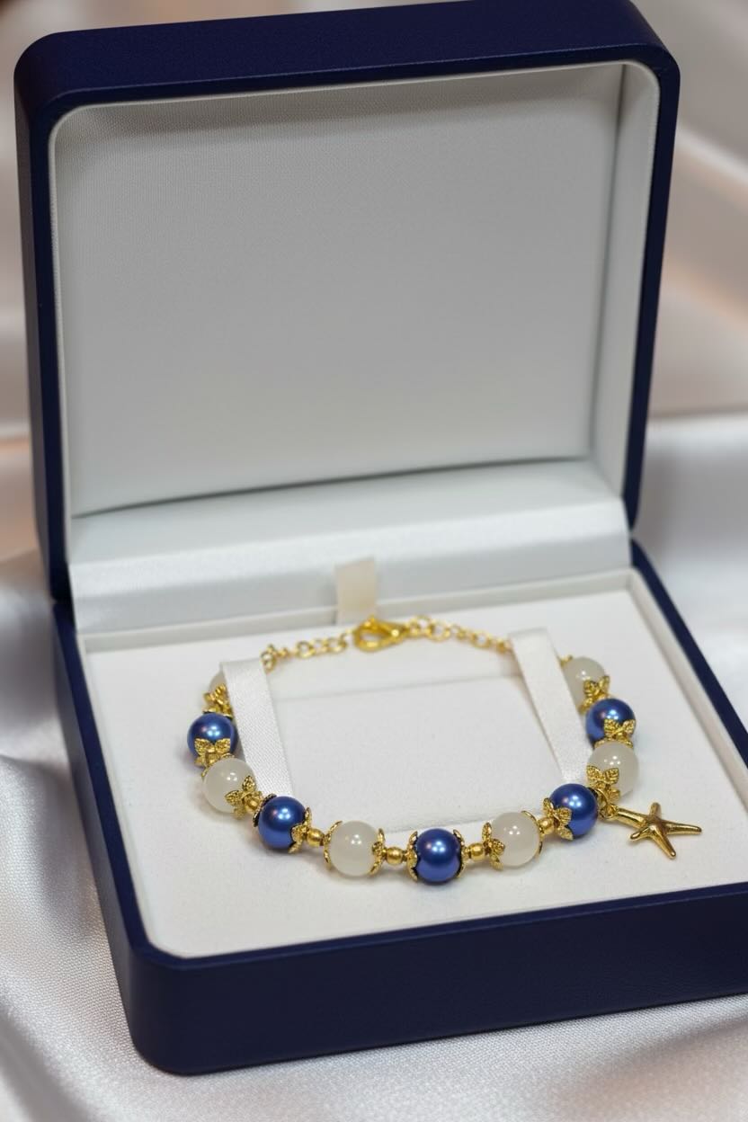 A gold-plated beaded bracelet with blue and white beads, displayed inside an open navy blue jewelry box on a white satin background.