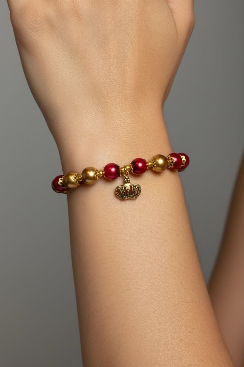 A close-up of a person's wrist wearing a beaded bracelet with red and gold glass pearls and a small crown charm, set against a neutral gray background.