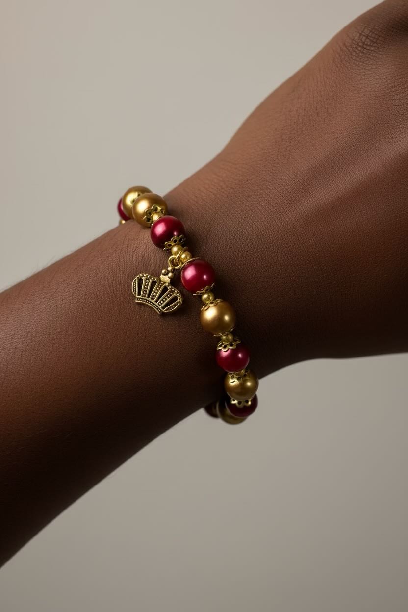 A close-up of a person's wrist wearing a beaded stretch bracelet with alternating gold and red pearls, accented by a small gold crown charm.