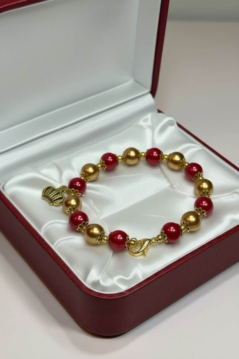 A beaded bracelet with glossy red and metallic gold glass pearls, featuring a small gold heart and crown charm and a lobster clasp, displayed inside an open red presentation box with a white satin lining.