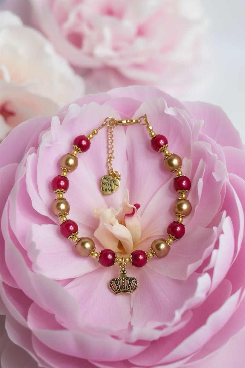 A gold-tone beaded bracelet with alternating deep red and and gold glass pearls, featuring a crown charm and a small heart charm, displayed on a large pink flower.