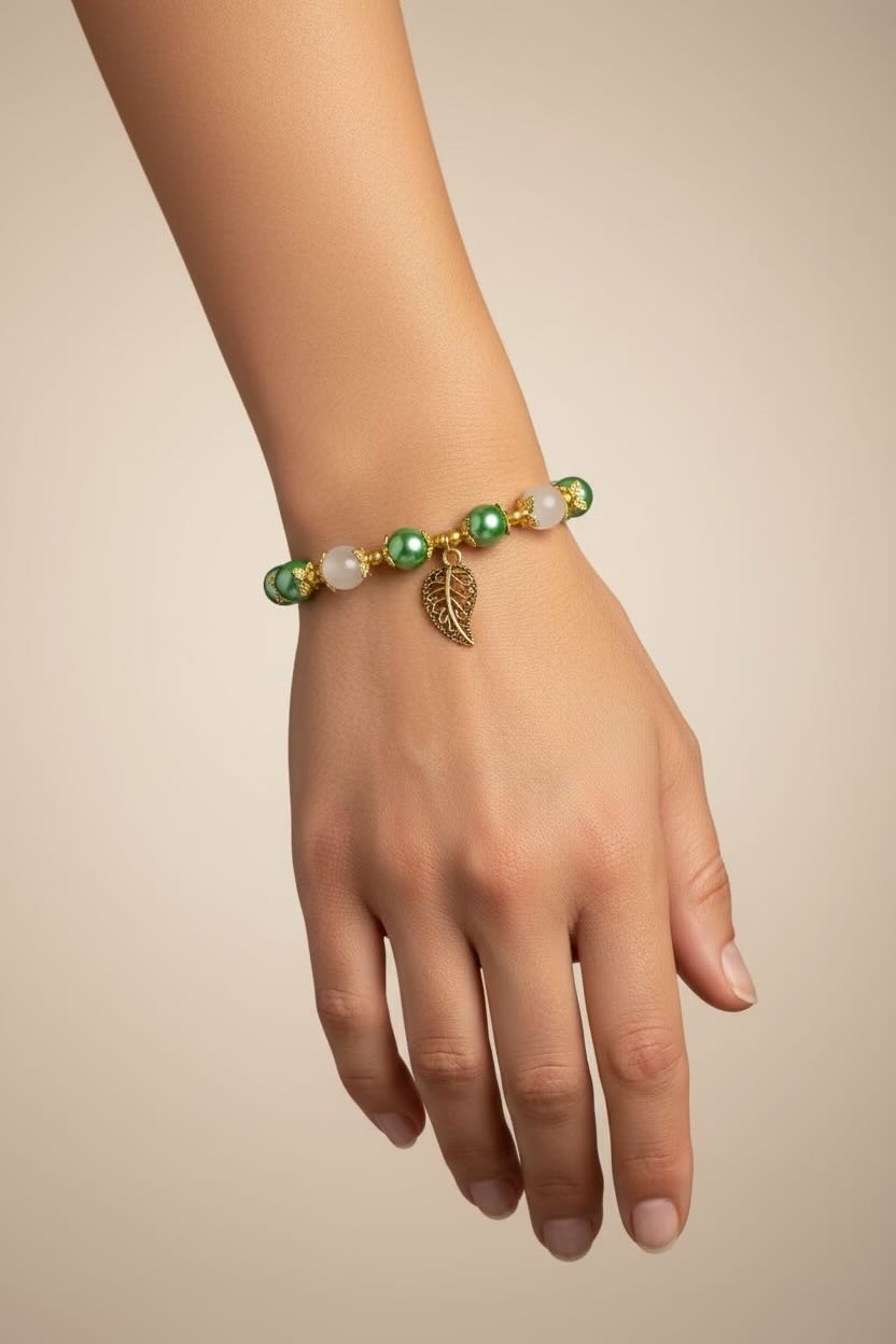 A close-up of a person's hand and wrist wearing a beaded bracelet with green and white glass pearls, gold-toned accents, and a dangling leaf charm, set against a plain beige background.