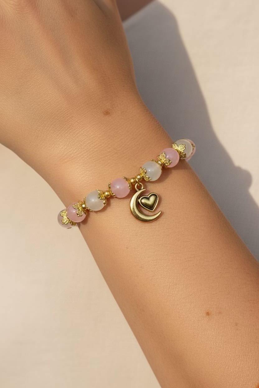 A close-up of a person's wrist wearing a delicate beaded bracelet with pink and white beads, gold-toned spacers, and a crescent moon charm with an inset heart.