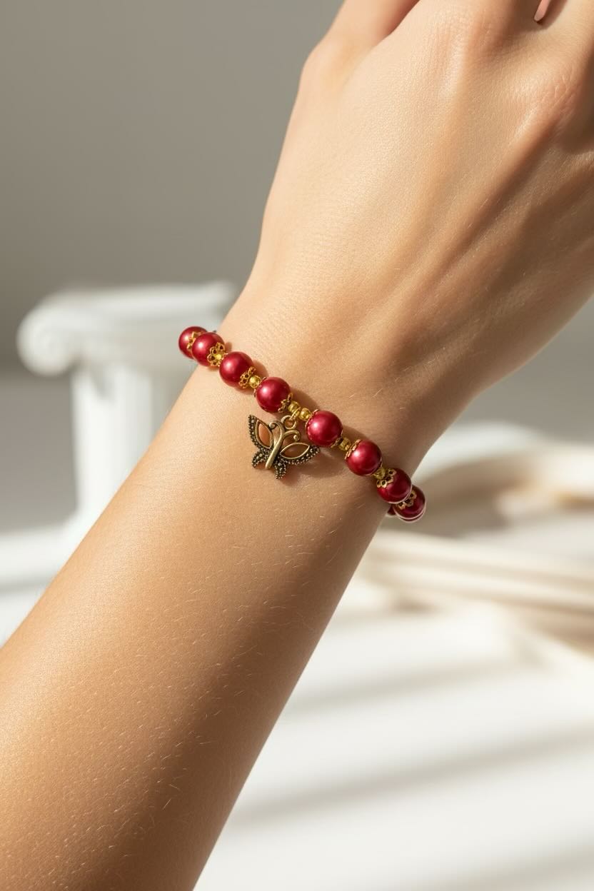 A close-up of a person's wrist wearing a beaded bracelet with red and black glass pearls, gold spacer details, and a small gold butterfly charm.