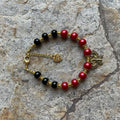 A beaded bracelet with alternating red and black glass pearls, gold-toned spacer beads, a gold clasp, a small heart charm, and a decorative metal butterfly piece, photographed on a gray stone surface.