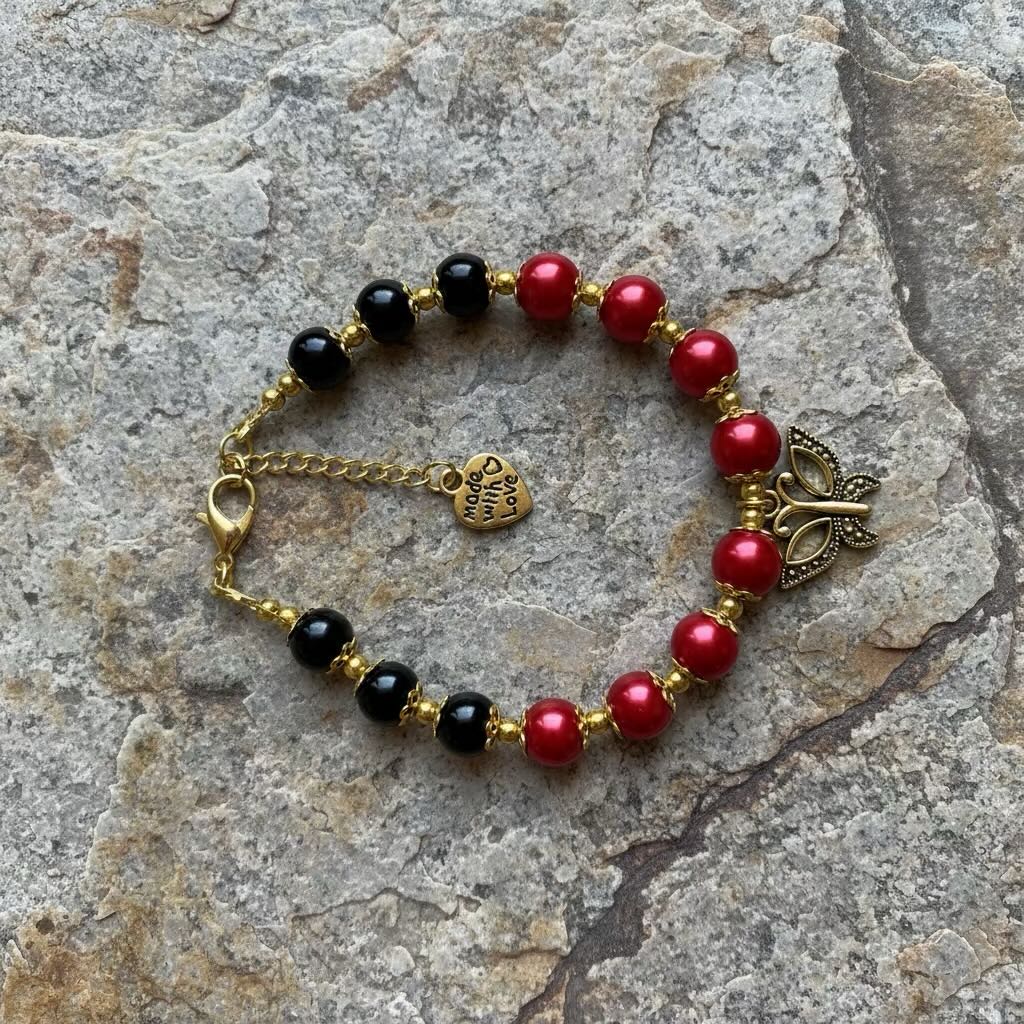 A beaded bracelet with alternating red and black glass pearls, gold-toned spacer beads, a gold clasp, a small heart charm, and a decorative metal butterfly piece, photographed on a gray stone surface.
