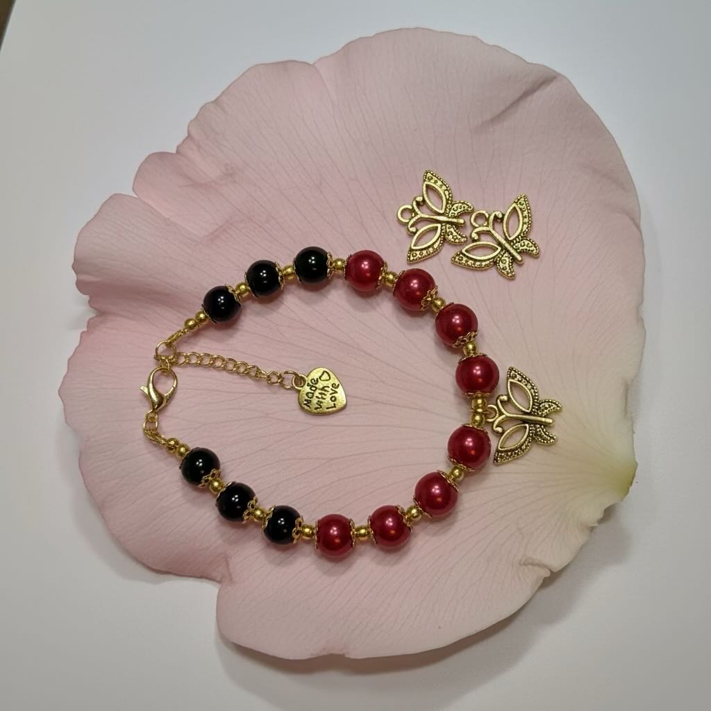 A beaded bracelet with alternating glossy red and black glass pearls, accented by gold-tone butterfly charms and a small heart-shaped tag, displayed on a large pink flower petal.